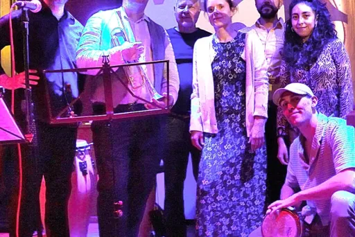 Gruppe von sieben Menschen auf einer Bühne, bunt beleuchtet, zwei halten Trommeln.