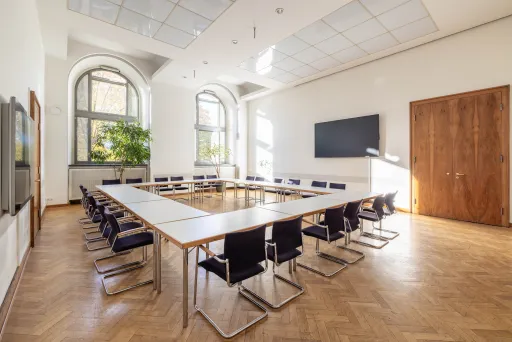 Heller Konferenzraum mit U-förmigem Tisch, Stühlen und großem Bildschirm an der Wand.