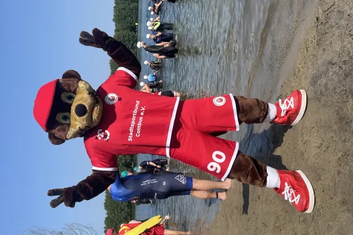 Maskottchen im roten Sportoutfit steht am Seeufer, Menschen im Wasser im Hintergrund.