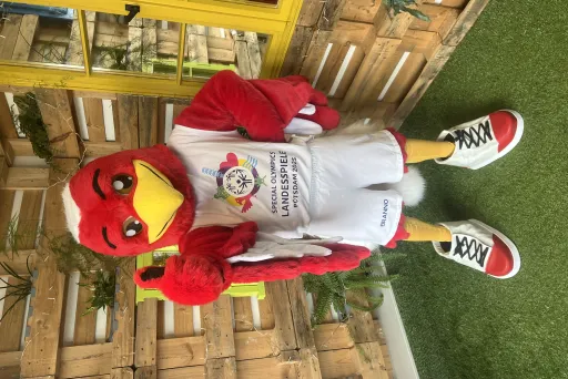 Maskottchen in Vogelkostüm mit weißem T-Shirt und Shorts auf Kunstrasen vor Holzwand.