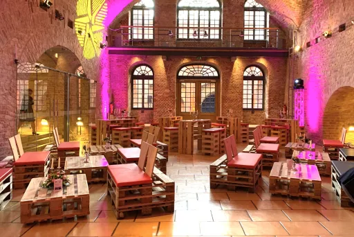 Gemütliche Lounge mit Palettenmöbeln und roten Kissen, Backsteinwände, pinke Beleuchtung.