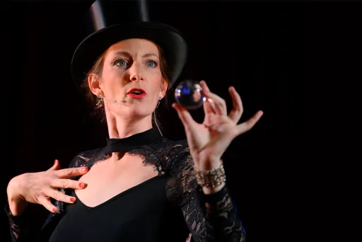 Frau mit Zylinder und schwarzem Kleid hält eine Kugel in der Hand auf dunklem Hintergrund.