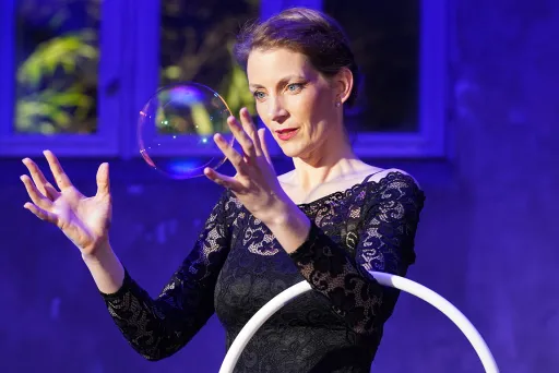 Frau in schwarzem Kleid hält Seifenblase in der Luft, lila Hintergrund