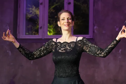 Frau im schwarzen Spitzenkleid lächelt und gestikuliert vor lila beleuchteter Wand.