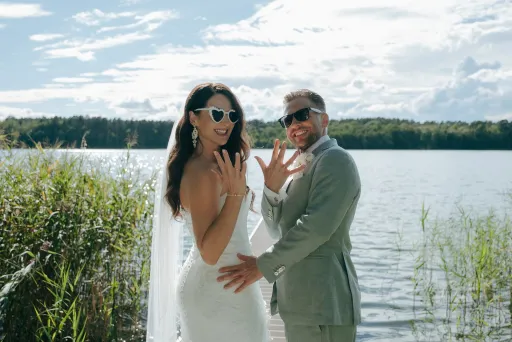 Brautpaar mit Sonnenbrillen zeigt stolz die Eheringe am Seeufer.
