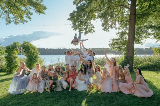 Gruppe fröhlicher Menschen in festlicher Kleidung posiert im Grünen am See, Baum im Hintergrund.