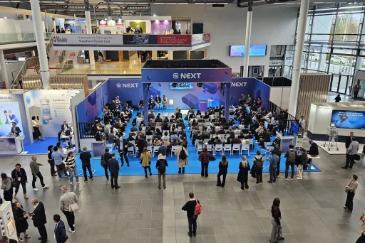 Viele Menschen sitzen und stehen vor einer Bühne auf einer Messe.