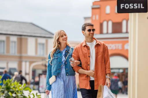 Paar spaziert lächelnd mit Einkaufstaschen durch eine Einkaufsstraße bei bewölktem Himmel.