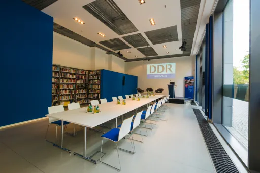 Moderner Konferenzraum mit langer Tafel, blau-weißen Stühlen und Bücherregalen.