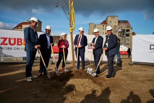 Sechs Personen mit Helmen pflanzen gemeinsam einen Baum auf einer Baustelle.