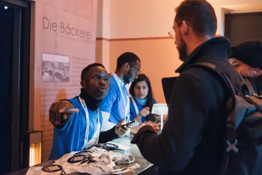 Ein Mann am Empfangstisch spricht mit einem Besucher, mehrere Personen im Hintergrund.