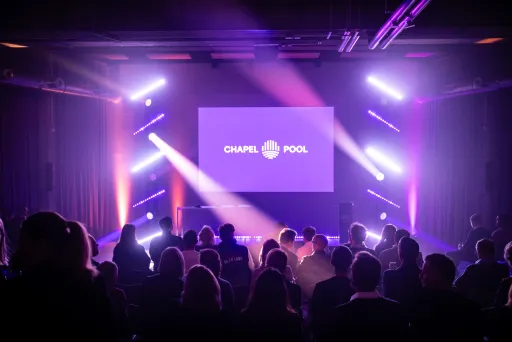 Publikum sitzt vor einer beleuchteten Bühne mit lila Licht und großem Bildschirm.