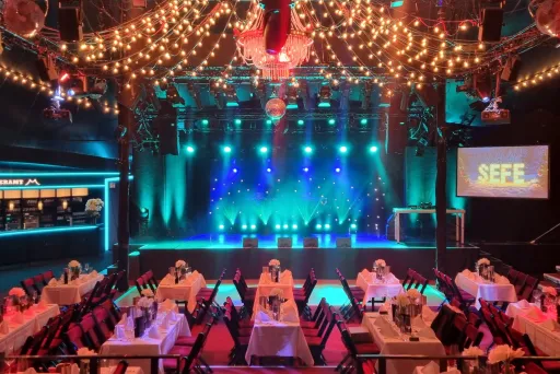 Festlich dekorierter Veranstaltungssaal mit gedeckten Tischen und bunter Bühnenbeleuchtung.