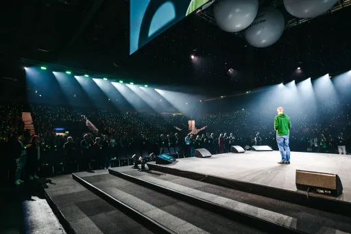 Person auf Bühne spricht vor großem, beleuchtetem Publikum in Arena.