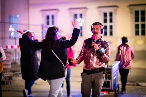 Moderator mit Mikrofon und Funkkopfhörern begleitet Teilnehmende einer Silent Disco Tour, die ebenfalls Kopfhörer tragen und im urbanen Außenraum interaktiv teilnehmen.