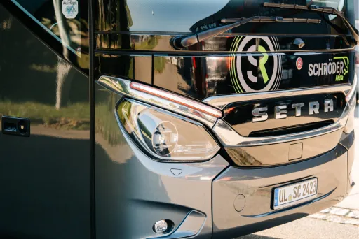 (2)	Nahaufnahme der Front eines modernen grauen Reisebusses von „Schröder Reisen“ mit Setra-Logo, Scheinwerfern und Kennzeichen.