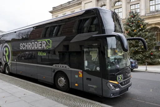 (3)	Grauer Doppeldecker-Reisebus von „Schröder Reisen“ steht am Straßenrand vor einem historischen Gebäude; daneben ist ein geschmückter Weihnachtsbaum zu sehen.