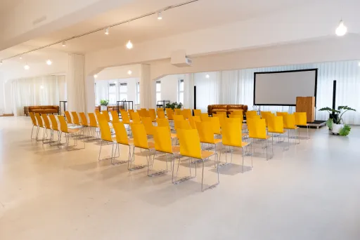 Heller Konferenzraum mit gelben Stühlen, Beamer und Leinwand.