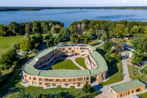 Modernes, ovales Gebäude mit begrüntem Dach am See, von Bäumen umgeben.