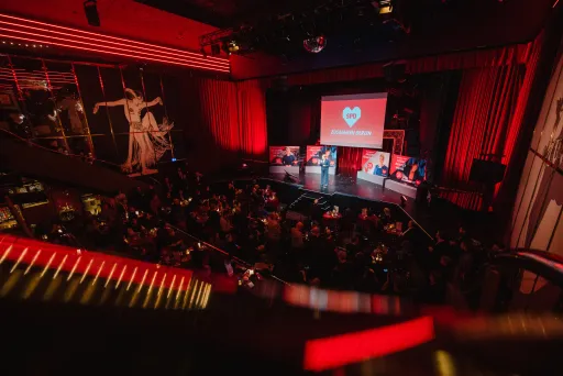Bühne mit rotem Licht, Publikum sitzt, Herzsymbol auf Leinwand im Hintergrund.