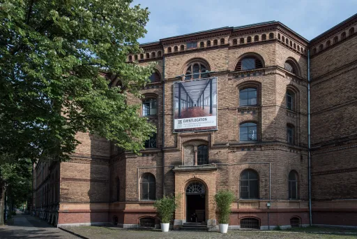 Backsteingebäude mit großem Werbebanner am Eingang, davor zwei Pflanzen.