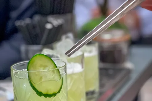 Glas mit gelbem Cocktail, Gurkenscheibe und Strohhalm auf Bartresen.