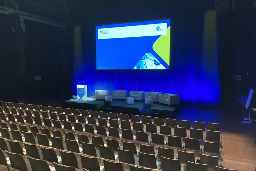 Leerer Konferenzsaal mit Bühne, Sofas und großem Bildschirm, blau beleuchtet.