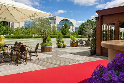 Sonnige Terrasse mit roten Teppich, Sitzbereich, Blumen und grüner Umgebung, Kanzleramt im Hintergrund.