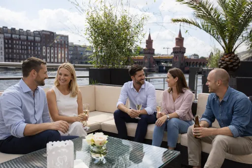 Fünf Menschen sitzen lachend auf einer Terrasse mit Blick auf die Oberbaumbrücke.