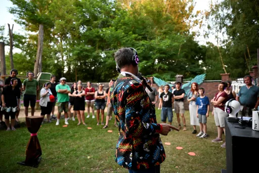 Moderator mit Mikrofon und Kopfhörern leitet ein interaktives Quiz im Freien; die Teilnehmenden stehen im Kreis und tragen kabellose Kopfhörer.
