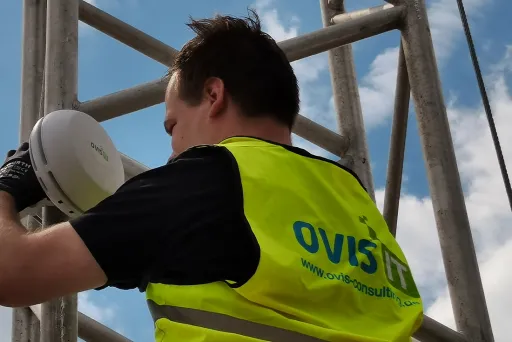Techniker mit gelber Weste montiert Gerät an Metallgerüst bei blauem Himmel.