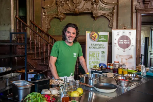 Mann mit langen Haaren in grüner Küche, lächelnd hinter einer Kochstation mit Zutaten.