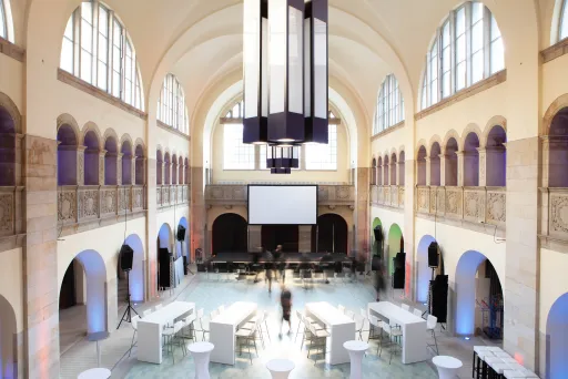 Großer, heller Veranstaltungssaal mit hohen Decken, Stehtischen und Leinwand.