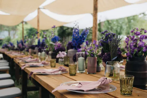 Lange Holztische mit lila Blumen und Gedecken im Freien, unter Sonnensegeln.