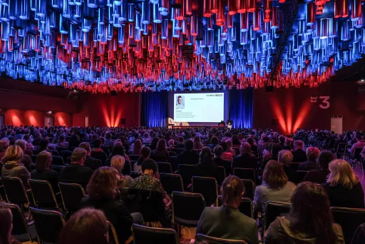 Großer Konferenzsaal mit vielen Zuschauern, Bühne vorne, bunte Deckenbeleuchtung.