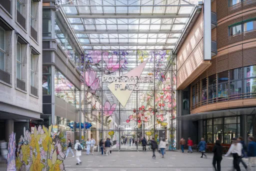 Überdachter Durchgang mit bunten Blumeninstallationen und vorbeigehenden Menschen.