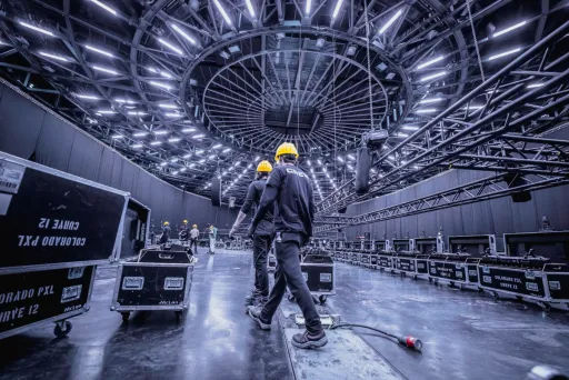 Zwei Arbeiter mit gelben Helmen unter einer futuristischen, beleuchteten Decke in einer Halle.