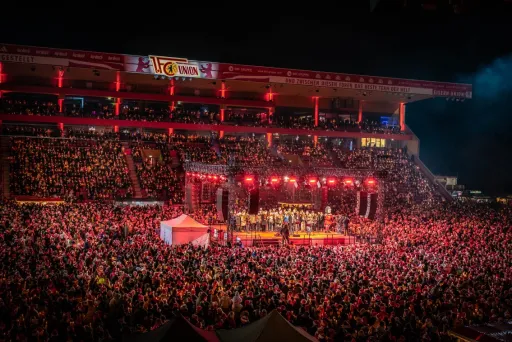 Große Menschenmenge bei einem Konzert im Stadion, Bühne leuchtet rot.