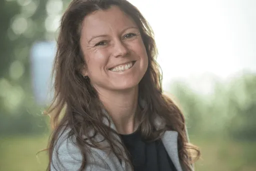 Lächelnde Frau mit langen braunen Haaren im Freien, unscharfer grüner Hintergrund.