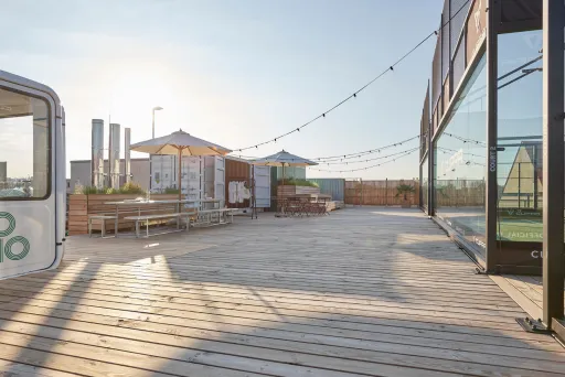 Sonnige Dachterrasse mit Holzboden, Tischen, Sonnenschirmen und Lichterketten.