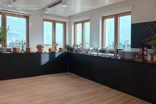 Heller Raum mit Holzboden, Fensterfront und schwarzer Theke, darauf Pflanzen und ein Cateringaufbau.