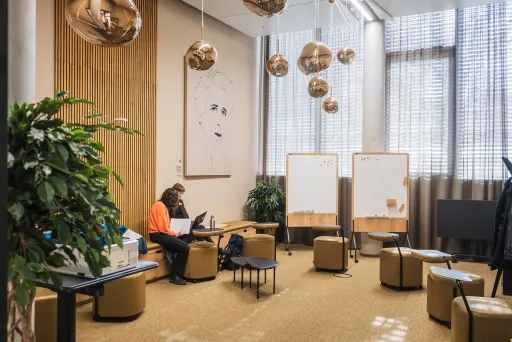 Frau arbeitet am Laptop in modernem, hellen Besprechungsraum mit Pflanzen und Sitzhockern.