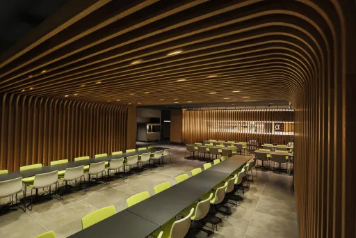 Modernes Restaurant mit langen Tischen, grünen Stühlen und Holzdecke.