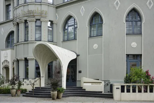 Gebäude mit rundem Vordach, großen Fenstern und Blumenkübeln am Eingang.