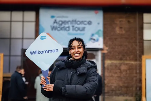 Transferhostess von cinnamon in schwarzer Winterjacke begrüßt Gäste lächelnd mit einem Willkommensschild bei der Ankunft.