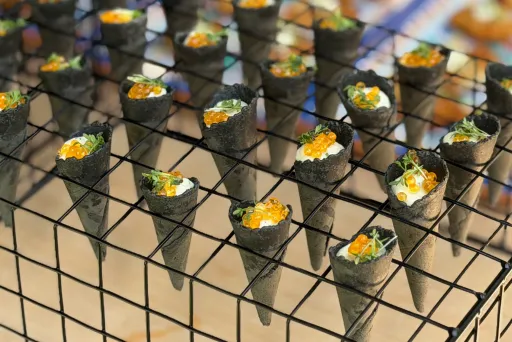Mehrere kleine Sushi-Handrollen mit Lachsrogen auf einem Metallgitter angerichtet.