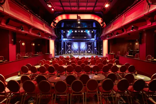 Rot beleuchteter Theatersaal mit runden Tischen und Stühlen, Blick auf die Bühne.