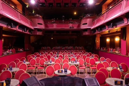 Leerer Theatersaal mit roten Stühlen und runden Tischen, Bühne im Vordergrund.
