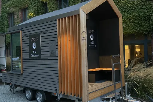 Modernes Tiny House auf einem Anhänger, umgeben von begrünten Gebäuden.
