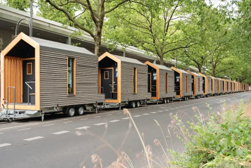 Reihe moderner Tiny Houses auf Anhängern unter Bäumen an einer Straße.